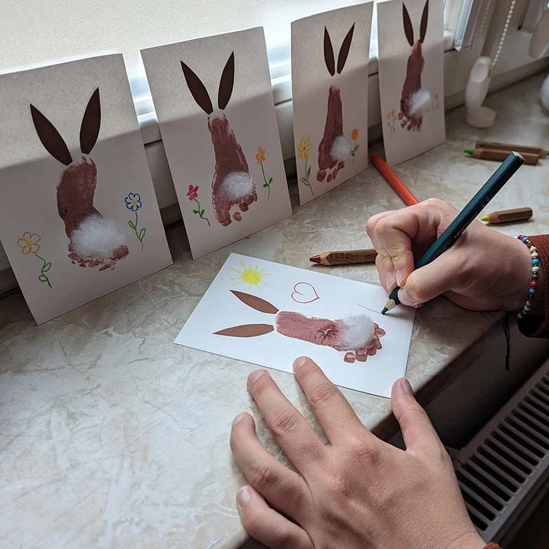 Eine Person verziert mit einem Stift eine Karte auf der aus einem Kinderfußabdruck bereits ein Osterhase gemalt wurde · Abenteuer Bindung · Sandra Bäthge in Magdeburg